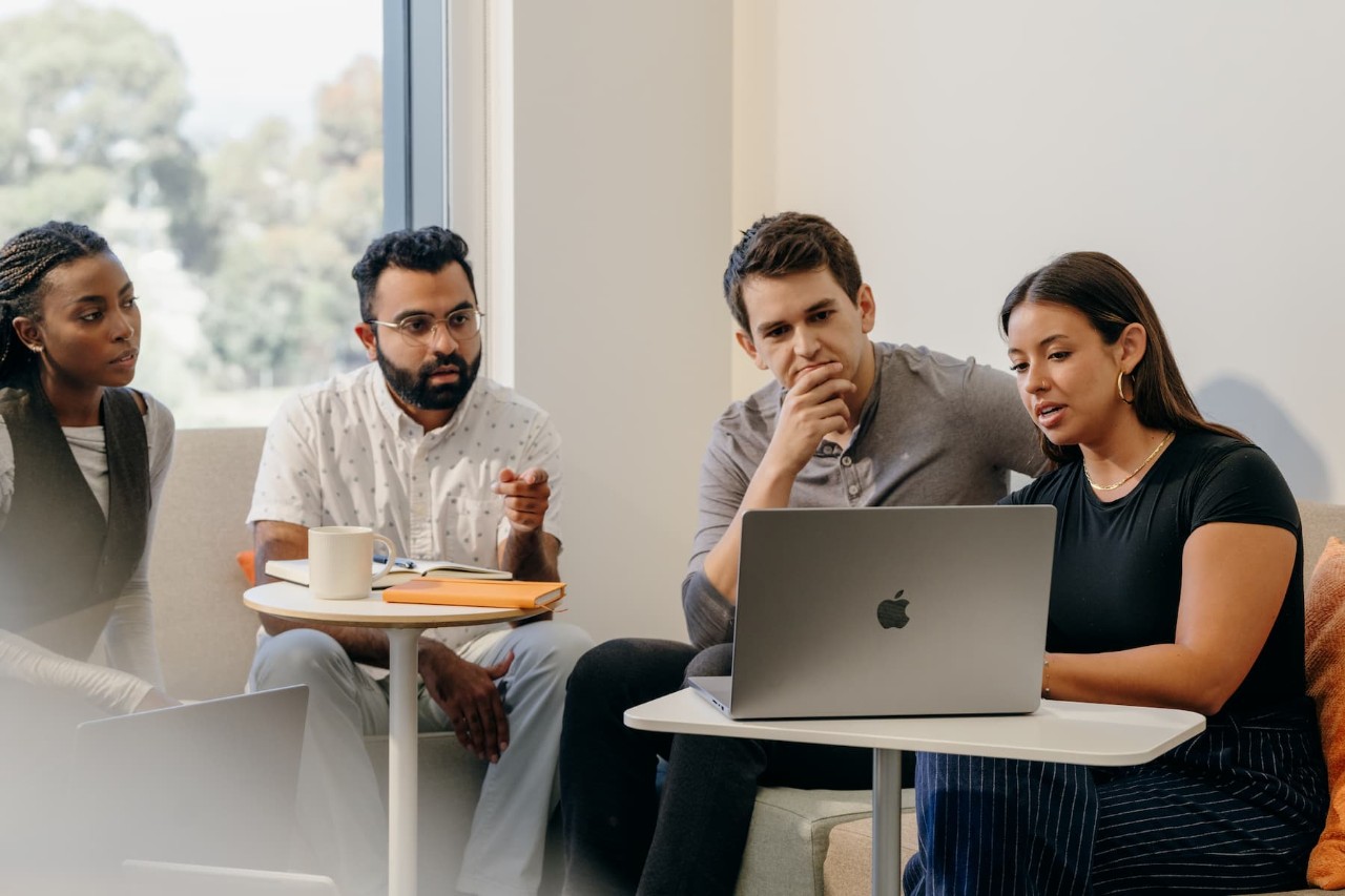 A group of diverse employees, two women and two men, are looking at an Apple laptop monitor, sitting in a casual office setting discussing and analyzing.