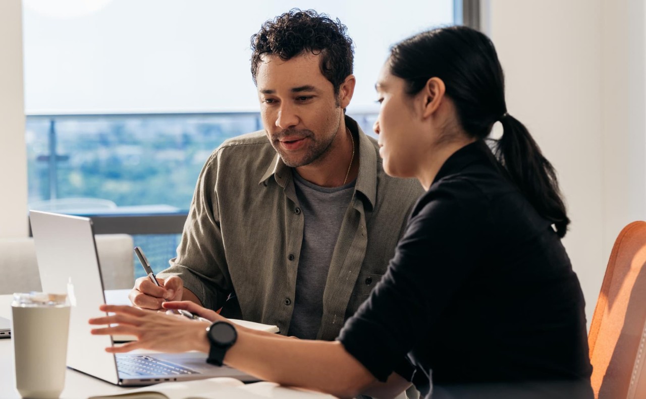 Two diverse employees are sitting at a table and looking at results on an Apple laptop, discussing and taking notes in notebooks.