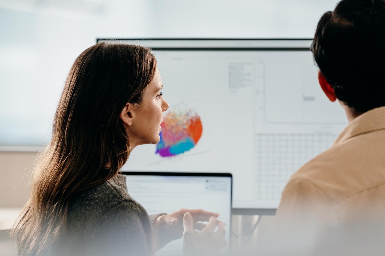 A close up, profile view of a woman and back view of a man in an office, analyzing and discussing data on dual screens.