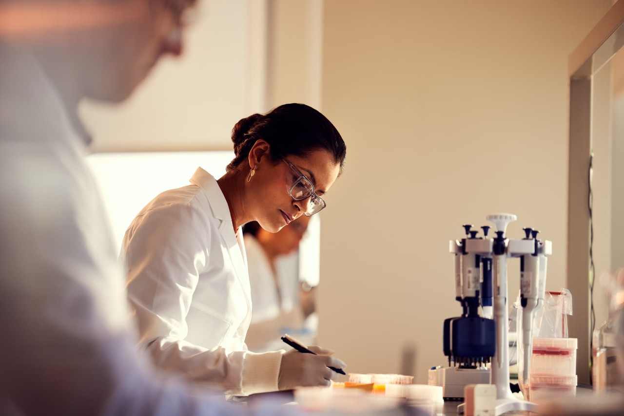 Female scientist, side view, writing information on lab bench, other scientists blurry in foreground and background