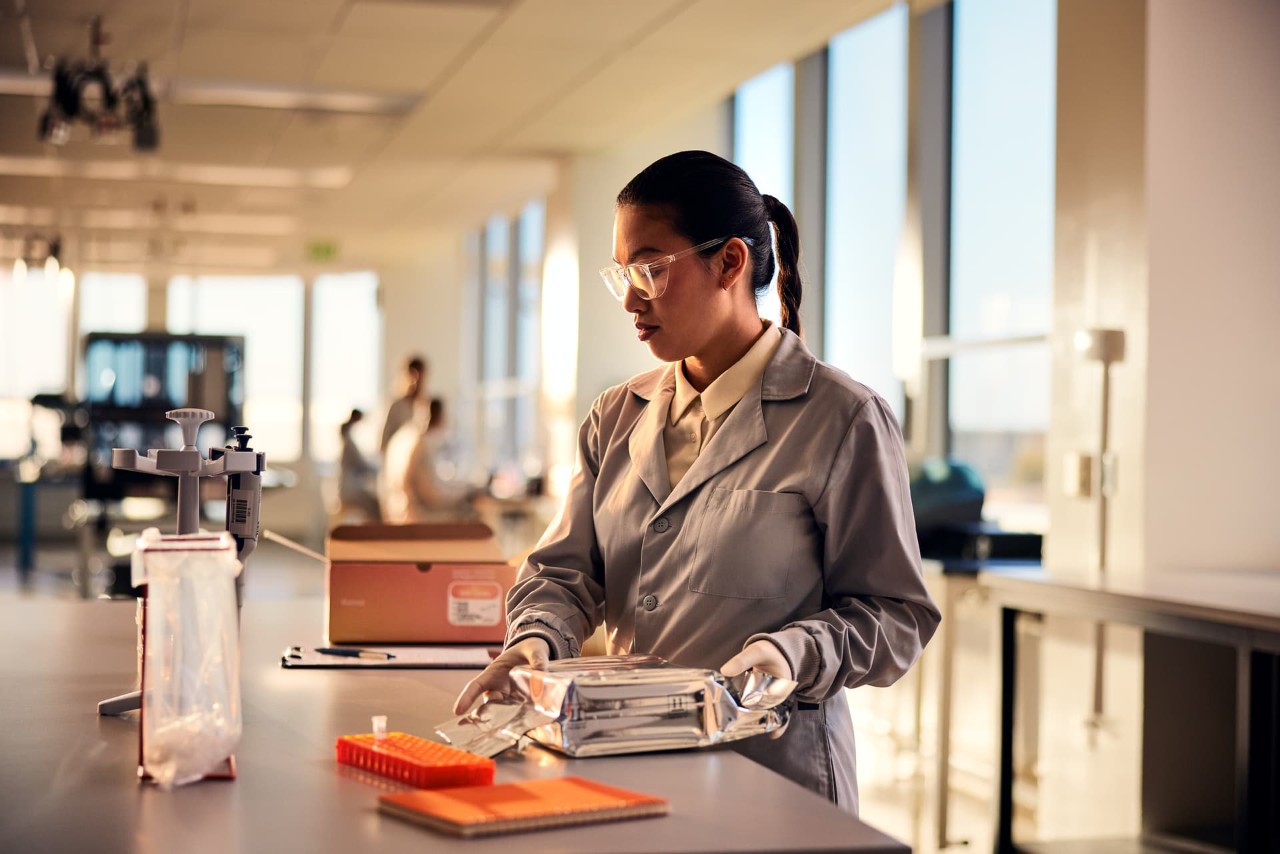 Female scientist, side view, unboxing NextSeq 1000/2000 cartridge with open P2 300 cycle box on lab bench, other scientists blurry in the background